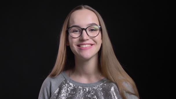 Primer plano de la joven atractiva hembra hipster en gafas mirando directamente a la cámara y sonriendo alegremente con el fondo aislado en negro — Vídeos de Stock