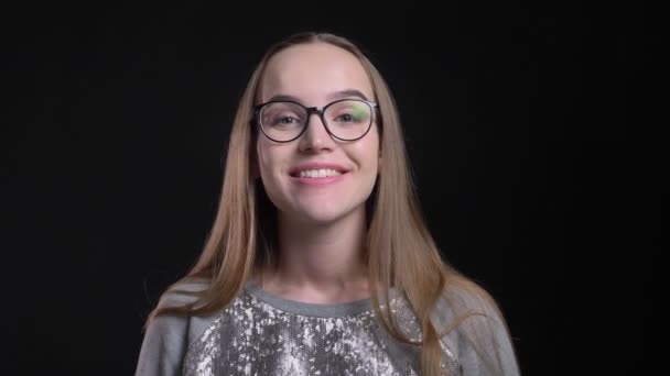 Gros plan tige de jeune hipster attrayant femelle dans des lunettes obtenir surpris et souriant regardant joyeusement droite à la caméra avec fond isolé sur noir — Video