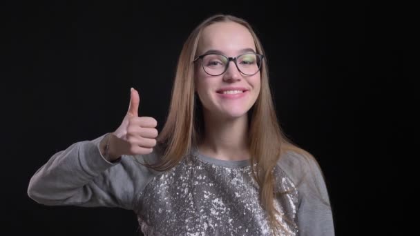 Closeup shoot of young attractive hipster female in glasses showing thumb up and smiling cheerfully while looking straight at camera with background isolated on black — Stock Video