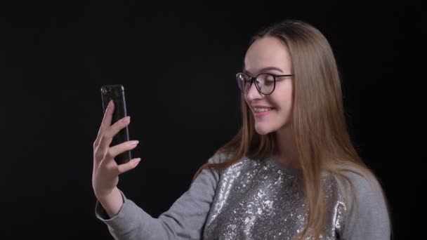 Съемки крупным планом молодой привлекательной хипстерки в очках с видеозвонком по телефону на фоне изолированного на черном фоне — стоковое видео