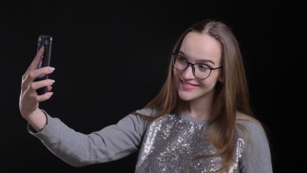 Vértes lő a fiatal, vonzó csípő női szemüveg selfies véve a telefon, és vidáman mosolyogva elszigetelt fekete háttérrel — Stock videók