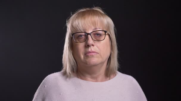 Съемки крупным планом старой белокурой женщины в очках, смотрящей прямо в камеру на фоне изолированного на черном фоне — стоковое видео