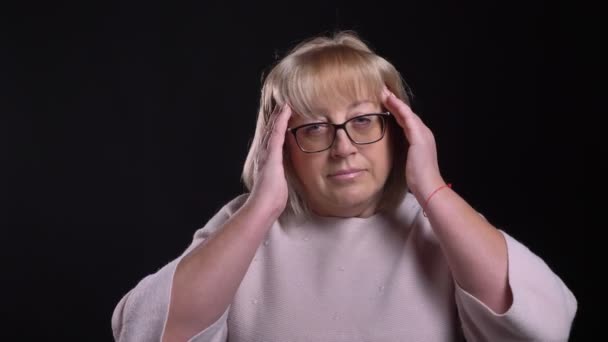 頭痛と背景が黒に分離されて疲れているメガネでシニアの白人金髪女性のクローズ アップ撮影 — ストック動画