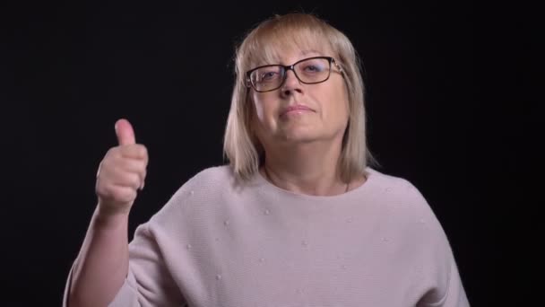 Съемка крупным планом старой белокурой женщины в очках с большим пальцем, смотрящей прямо в камеру на фоне, изолированном на черном — стоковое видео