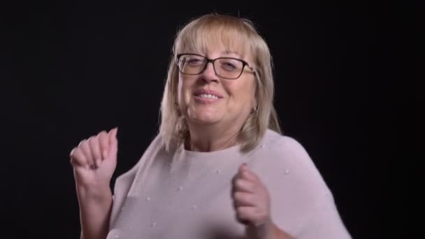 Eski Beyaz sarışın kadın dans ve having fun kamera önünde izole siyah arka plan ile gözlük closeups ateş — Stok video