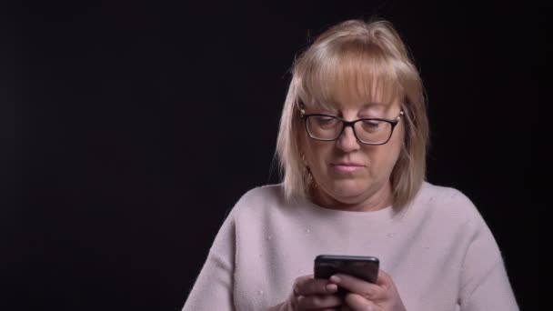 Close-up opnamen van Kaukasische blonde meisje in glazen de telefoon voor de camera gebruikt met geïsoleerd op zwarte achtergrond — Stockvideo