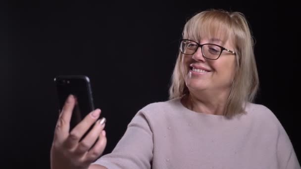 Primer plano de la vieja rubia caucásica en gafas tomando selfies en el teléfono delante de la cámara con fondo aislado en negro — Vídeos de Stock