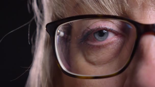 Primer plano de media cara de la cara femenina rubia caucásica senior en gafas con su ojo mirando directamente a la cámara con el fondo aislado en negro — Vídeo de stock