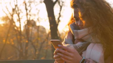 Smartphone ile memnun gülümsemek güneşli sonbahar Park içine izlerken bankta oturan kıvırcık saçlı beyaz kız portresi.