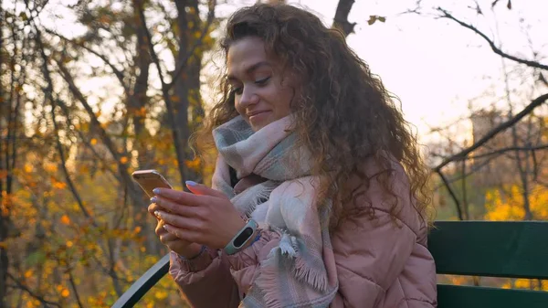 Porträt eines kaukasischen Mädchens mit lockigem Haar, das auf einer Bank sitzt und im herbstlichen Park genüsslich ins Smartphone schaut. — Stockfoto