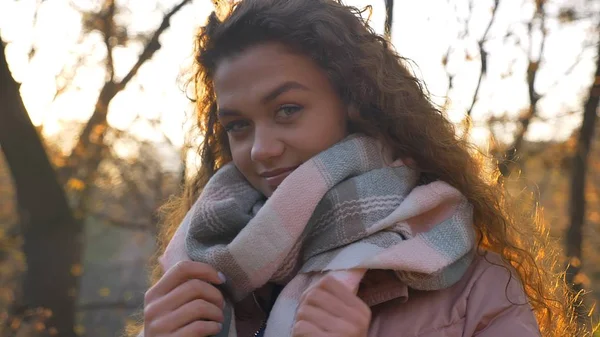 Porträtt av leende curly-haired kaukasiska flicka håller hennes halsduk och titta in i kameran i höstlig park. — Stockfoto