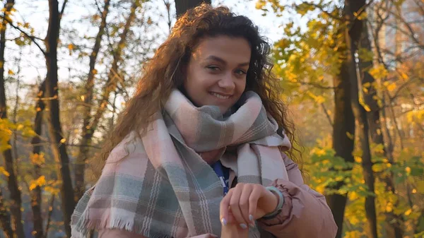 Porträtt av leende curly-haired kaukasiska flicka titta på klockor och in kameran i höstlig park. — Stockfoto