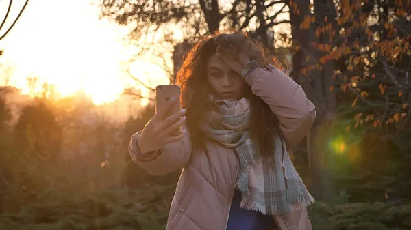 Retrato de menina caucasiana jovem ajustando seu cabelo e fazendo fotos de selfie usando smartphone no parque outonal . — Fotografia de Stock