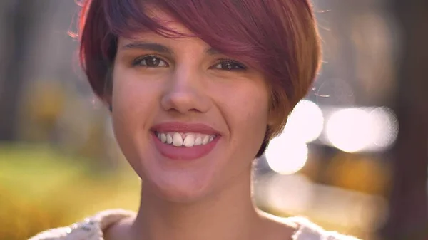 Close-up portret młodego fajny kaukaski haired Dziewczyna oglądania uśmiechem do aparatu na tle słoneczny park. — Zdjęcie stockowe