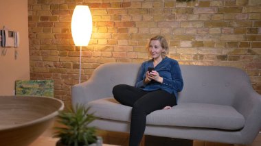 Smartphone ile kanepede oturan ve sola doğru rahat ev ortamında büyük bir ilgi ile izlerken üst düzey beyaz Bayan portresi.