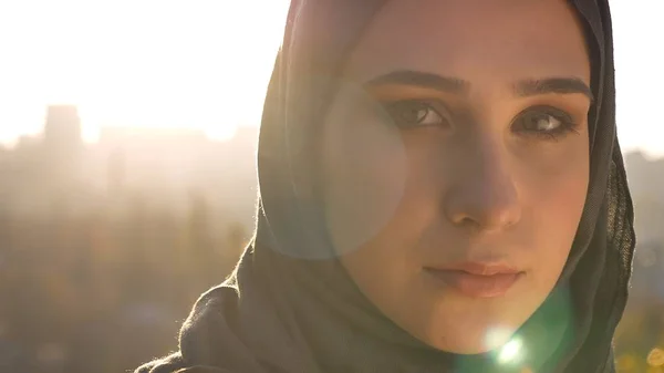 Closeup shoot of young attractive muslim female in hijab looking forward with urban setting and shining sun on the background.