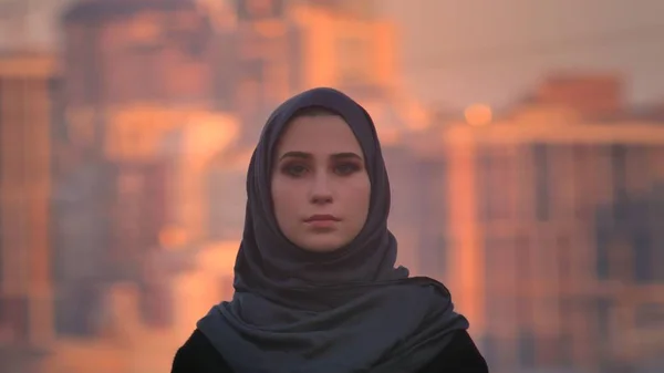 Primer plano retrato de joven atractiva hembra en hijab mirando directamente a la cámara con la ciudad urbana en el fondo —  Fotos de Stock