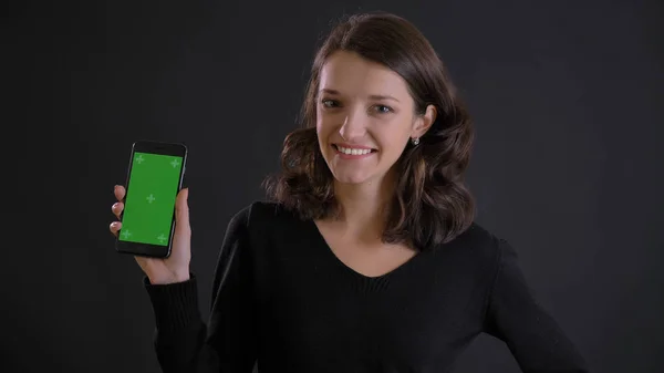 Портрет милої молодої брюнетки, що показує зелений екран смартфона радісно на чорному тлі . — стокове фото