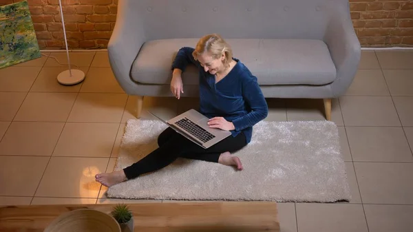 Portrait of senior caucasian lady sitting on floor and watching into laptop joyfully in cozy home atmosphere. — Stock Photo, Image