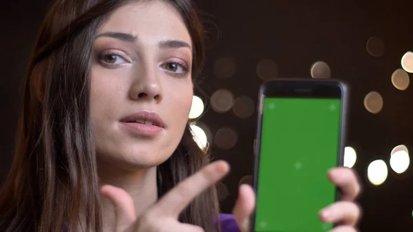 Portrait of beautiful caucasian brunette girl showing green screen of smartphone into camera on blurred lights background. — Stock Photo, Image