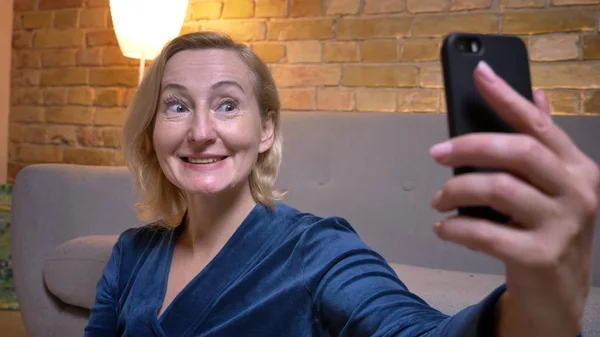 Alegre señora caucásica mayor sentada en el suelo y haciendo felices selfies-fotos utilizando el teléfono inteligente en ambiente acogedor en casa . —  Fotos de Stock