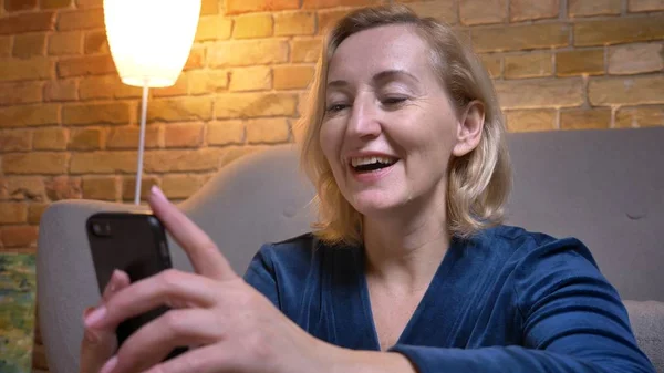 Nahaufnahme Porträt einer fröhlichen kaukasischen Seniorin, die ins Smartphone schaut und in gemütlicher Atmosphäre zu Hause lacht. — Stockfoto