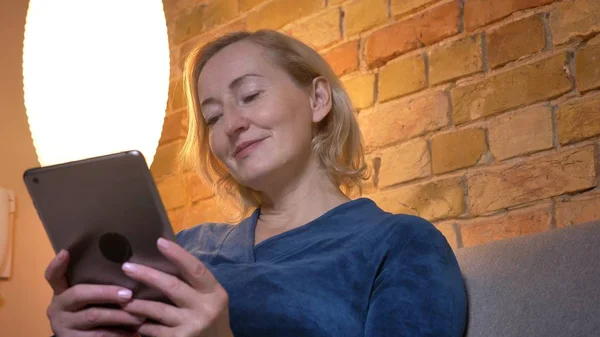 Close-up portret van senior Kaukasische dame zittend op de Bank en kijken naar tablet aandachtig en glimlachend in gezellige huis sfeer. — Stockfoto