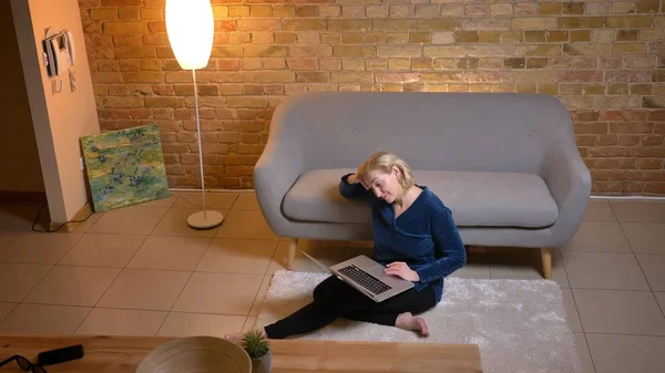 Portret van senior Kaukasische vrouw zittend op de vloer en kijken op laptop met amusement in gezellige huis sfeer. — Stockfoto