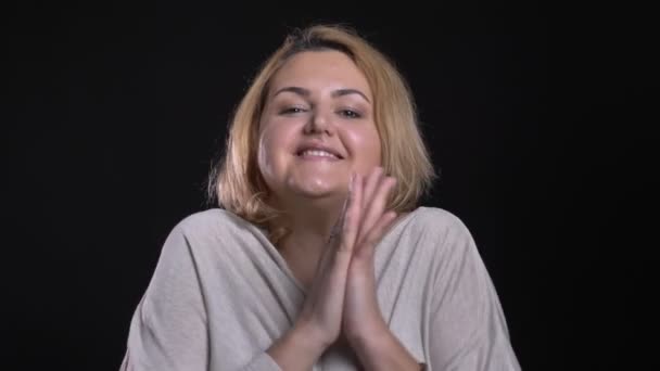 Blonde short-haired overweight businesswoman claps her hands showing gladness into camera on black background.