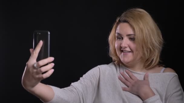 Cute blonde short-haired overweight businesswoman talking joyfully in videochat on smartphone on black background.