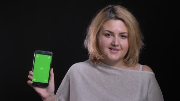 Blonde kurzhaarige übergewichtige Geschäftsfrau demonstriert grünen Bildschirm des Smartphones auf schwarzem Hintergrund. — Stockvideo