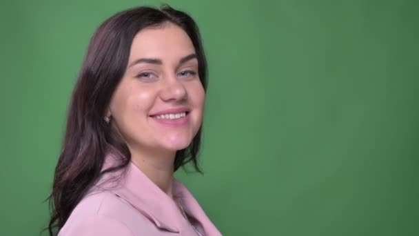 Young brunette businesswoman in pink jacket turns to camera and watches delightedly into camera on green background.