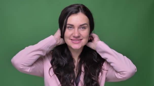 Mooie brunette zakenvrouw in roze jas tot vaststelling van haar haren en schuchter glimlachend in de camera op groene achtergrond. — Stockvideo