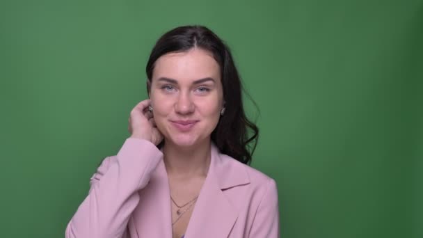 Schöne brünette Geschäftsfrau in pinkfarbener Jacke, die ihr Haar fixiert und bescheiden in die Kamera auf grünem Hintergrund lächelt. — Stockvideo