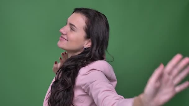 Portret van jonge brunette vrouwelijke vloger in roze jasje dansen met tevredenheid in camera op groene achtergrond. — Stockvideo