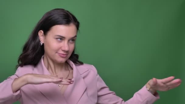 Портрет юной брюнетки-видеоблогера в розовой куртке странно танцующей и подмигивающей в камеру на зеленом фоне . — стоковое видео
