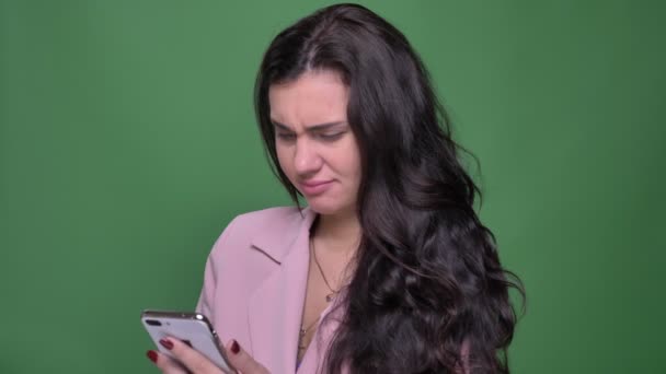 Portrait of young brunette female vloger in pink jacket watching into smartphone and emotionally reacting on green background. — Stock Video