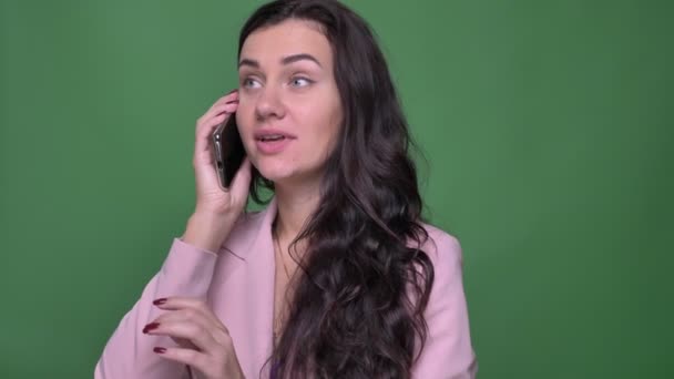 Portré, fiatal barna nő beszélgetett barát örömmel smartphone zöld háttér pink dzseki. — Stock videók