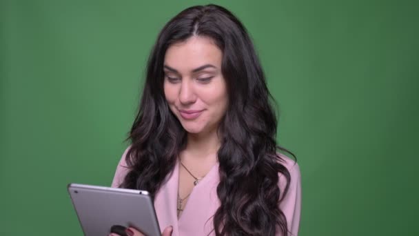 Retrato de la joven morena vloger femenina en chaqueta rosa mirando sonriente en la tableta sobre fondo verde . — Vídeos de Stock