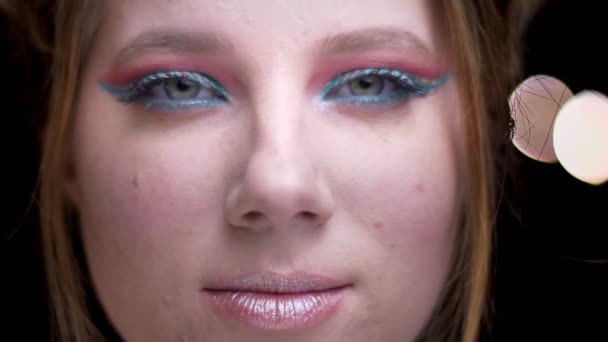 Porträt eines kaukasischen blonden Mädchens mit leuchtend buntem Make-up, das friedlich in die Kamera auf verschwommenem Licht blickt. — Stockvideo