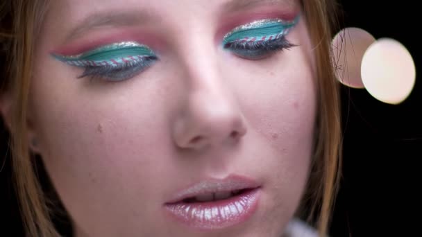 Menina loira com maquiagem multicolorida virando à esquerda e assistindo misteriosamente em câmera em luzes borradas fundo . — Vídeo de Stock