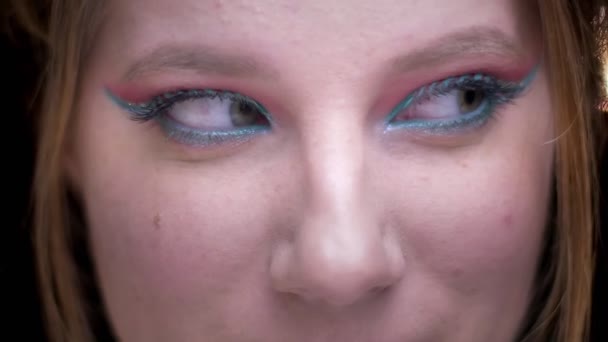 Close-up portret van blonde model met kleurrijke make-up heen en weer bewegen van haar ogen op wazig lights achtergrond. — Stockvideo