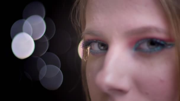 Close-up portrait of fair-haired model turns to camera and watches seriously slowly blinking on blurred lights background. — Stock Video