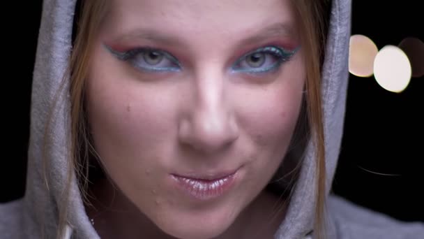 Porträt einer kaukasischen Blondine im Kapuzenpulli mit grell buntem Make-up, die auf verschwommenem Licht lächelt. — Stockvideo