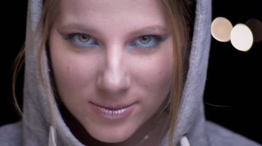 Sportsgirl gözleri utanarak yetiştirme ve kameraya gülme renkli makyaj ile hoodie içinde ışıklar arka plan bulanık.