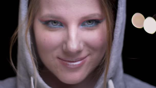 Blonde sportsgirl in hoodie met kleurrijke make-up schuchter glimlachend in de camera op wazig lights achtergrond. — Stockvideo