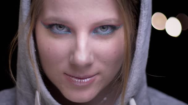Sportsgirl in hoodie met kleurrijke make-up verhoging van haar ogen schuchter en lachen in de camera op de achtergrond wazig lights. — Stockvideo