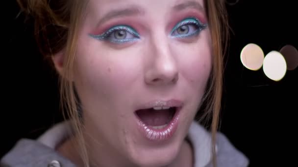 Nahaufnahme Porträt eines blonden Mädchens mit buntem Make-up, das positive Belustigung kindlich auf verschwommenem Licht-Hintergrund zeigt. — Stockvideo