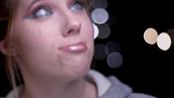 Retrato de cerca de una chica rubia con maquillaje colorido que sopla su cabello de la cara alegremente sobre un fondo borroso de luces . — Vídeo de stock