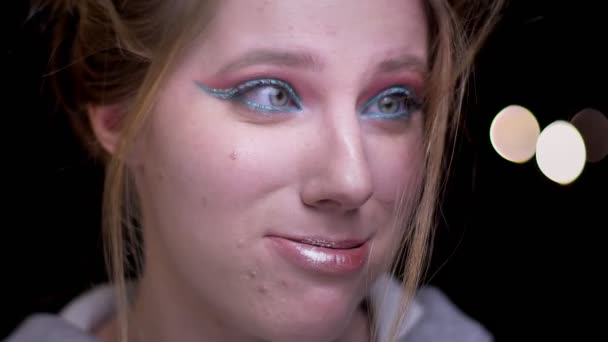 Retrato de chica rubia con maquillaje colorido soplando su cabello de la cara con frustración divertida en el fondo de luces borrosas . — Vídeo de stock